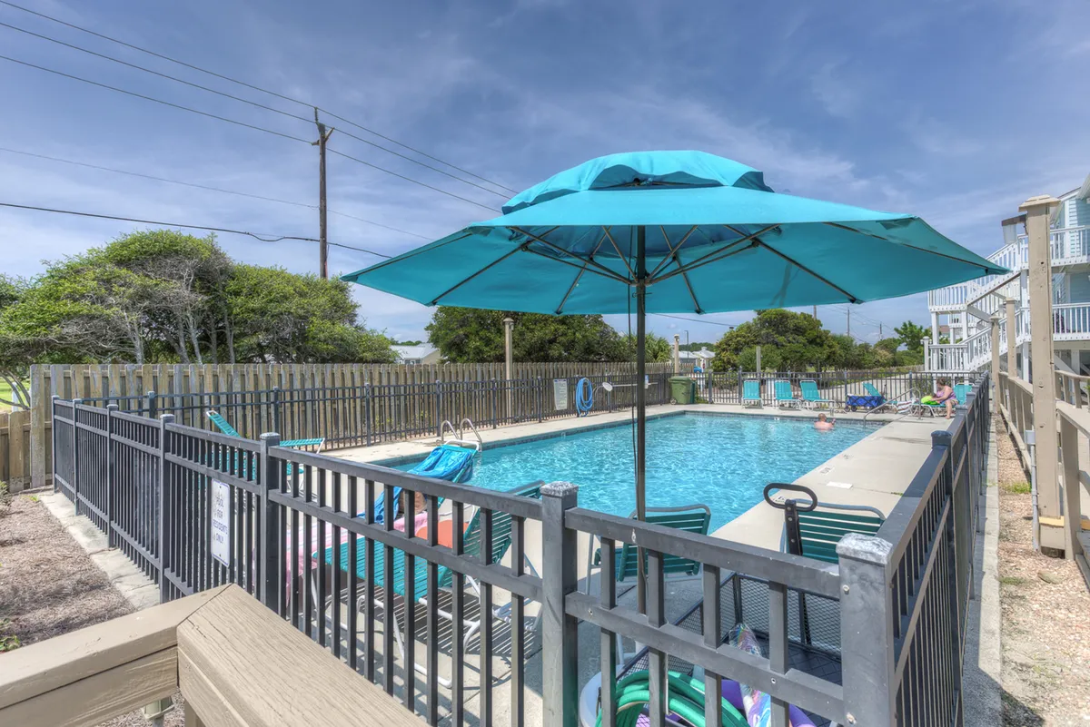 A vibrant outdoor pool scene with a large teal umbrella and inviting lounge chairs surrounded by lush greenery. The bright, sunlit setting suggests a lively, social atmosphere perfect for swimming, sunbathing, or gathering with friends for poolside fun.