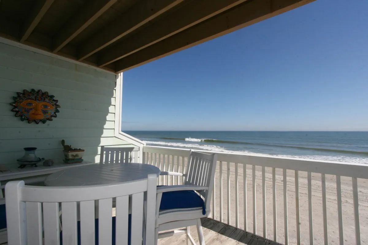 A serene patio seating area with comfortable chairs looks out over a beautiful stretch of beach and ocean, framed by a decorative sun mask on the wall. The horizon glows with soft light, signaling either sunrise or sunset, and the gentle waves are visible in the distance, inviting relaxation and oceanfront enjoyment.