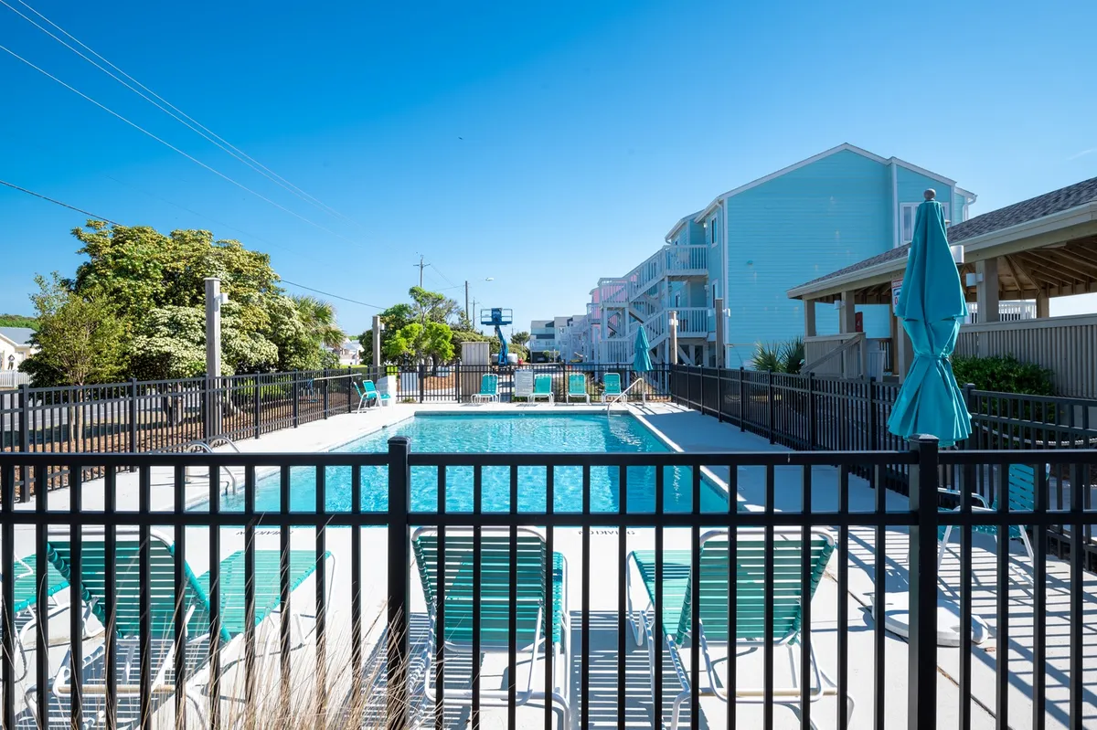 A bright, inviting scene featuring a large outdoor swimming pool surrounded by lounge chairs, umbrellas, and lush greenery. The atmosphere is leisurely, with visible spaces for sunbathing, playing, and socializing—perfect for families and children to enjoy summer days.
