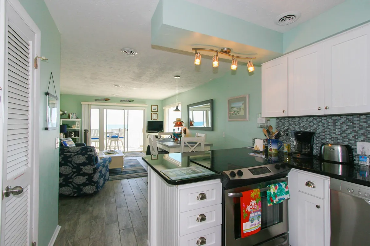 A sleek, modern kitchen with stainless appliances, a large island with breakfast bar seating, and an open floor plan that leads directly to a balcony or deck with ocean views. The space is filled with natural light and designed for both cooking and socializing.