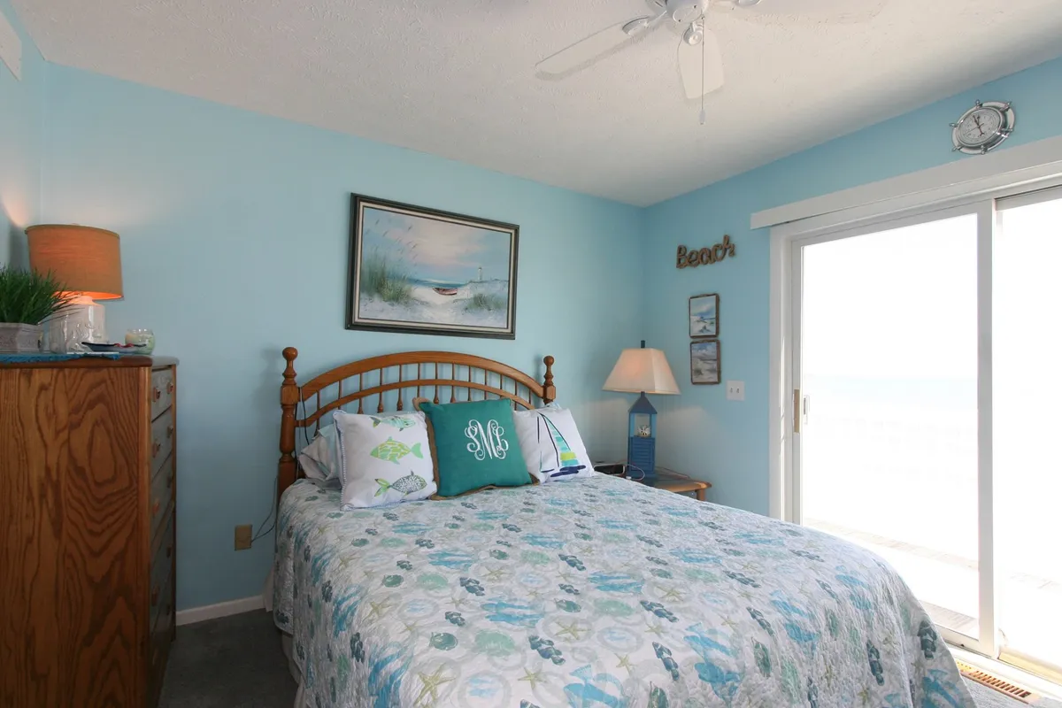 This image features a bedroom with a queen bed adorned with decorative pillows, tasteful wall art, and a sliding glass door that frames an inviting ocean view. The overall ambiance is calming, with natural light streaming in and an inviting deck just beyond the door, perfect for relaxing with a book or savoring the breeze.