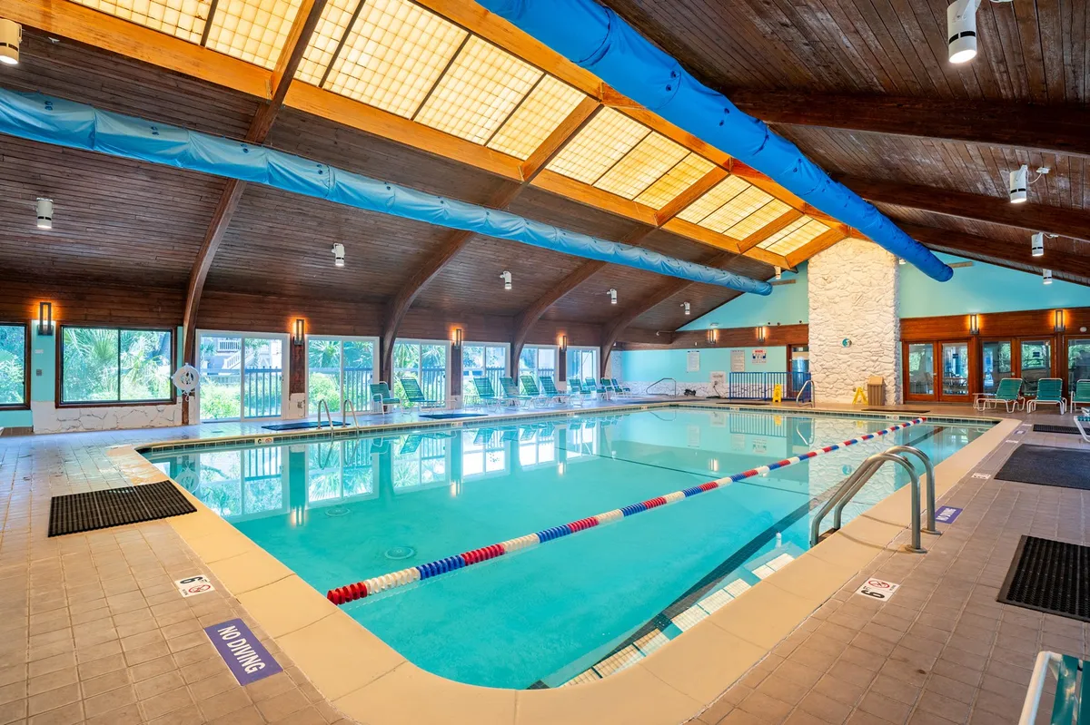 A tranquil indoor pool area featuring a wooden ceiling, large windows letting in natural light, and comfortable poolside lounge chairs. The space feels calm and perfect for relaxation or a swim, regardless of the weather.