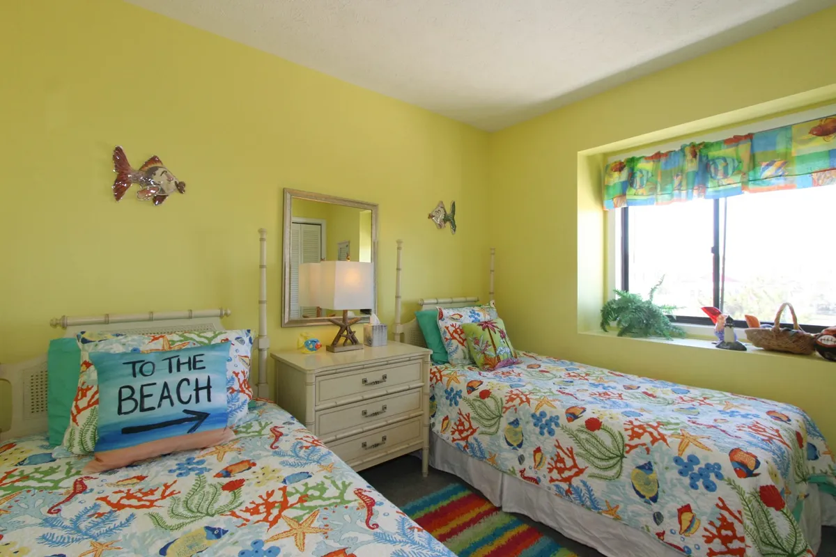 A cheerful second bedroom is shown with colorful beach-themed bedding, bright yellow walls, decorative fish accents, a mirror reflecting natural light, and a cozy nightstand lamp. The playful decor and welcoming space create a fun environment ideal for kids or friends.