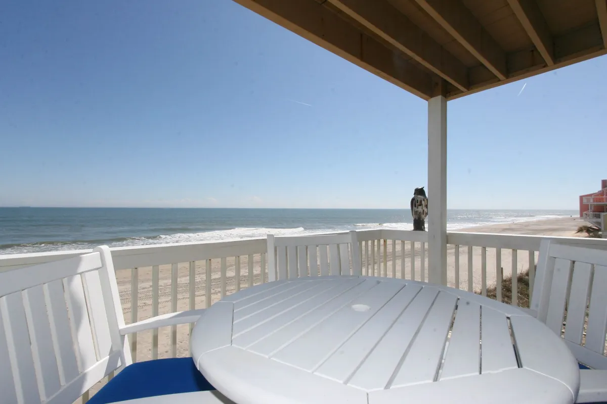 The image showcases a stunning oceanfront view from the condo's private balcony, featuring comfortable outdoor seating and a table—perfect for unwinding, enjoying meals, or soaking in the tranquil atmosphere. The endless blue of the Atlantic stretches beyond, with the golden light hinting at a serene sunrise or sunset.