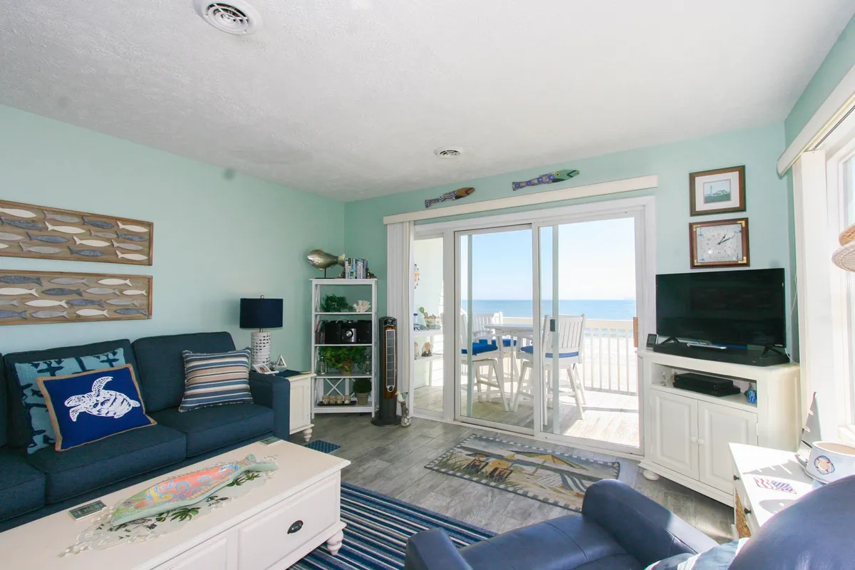 A bright and spacious open living area featuring cozy seating, a queen sleeper sofa, ocean-inspired décor, and large sliding glass doors framing expansive ocean views. The decorative fish art and cheerful ambiance invite relaxation and social gatherings.